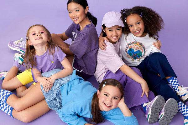 5 young girls huddled together, smiling at the camera in an array of purple, blue, yellow and white pieces from the Y2K Collegiate collection