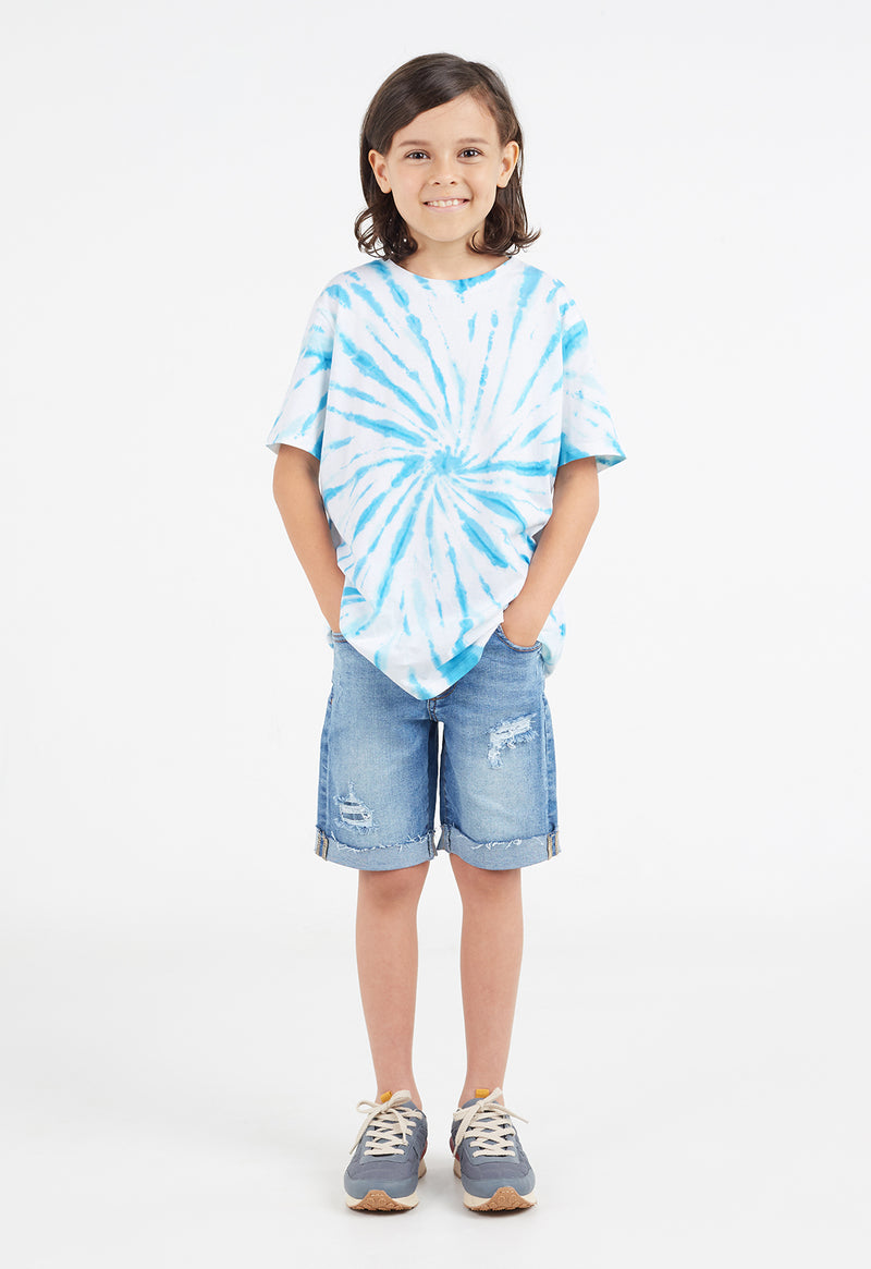 The young boy wears the Boys Blue and White Tie-Dye T-Shirt by Gen Woo with denim shorts and sneakers
