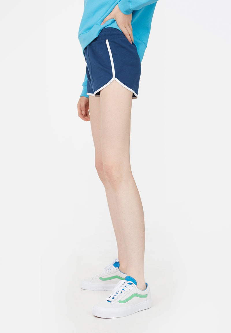 The model wears the Blue and White Retro Ladies Track Shorts by Gen Woo with trainers