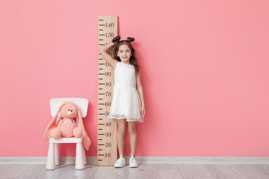 Rather than buying endless ill-fitting kids' clothes online, shop for your child's correct size by understanding kids’ size charts and conversions. A young girl measuring herself against a pink wall. (Adobe Stock / Pixel-Shot)