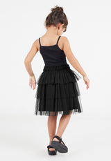 Back view of the young girl wearing the Black Mesh Tiered Knee-Length Girls Skirt by Gen Woo with a black cami and sandals
