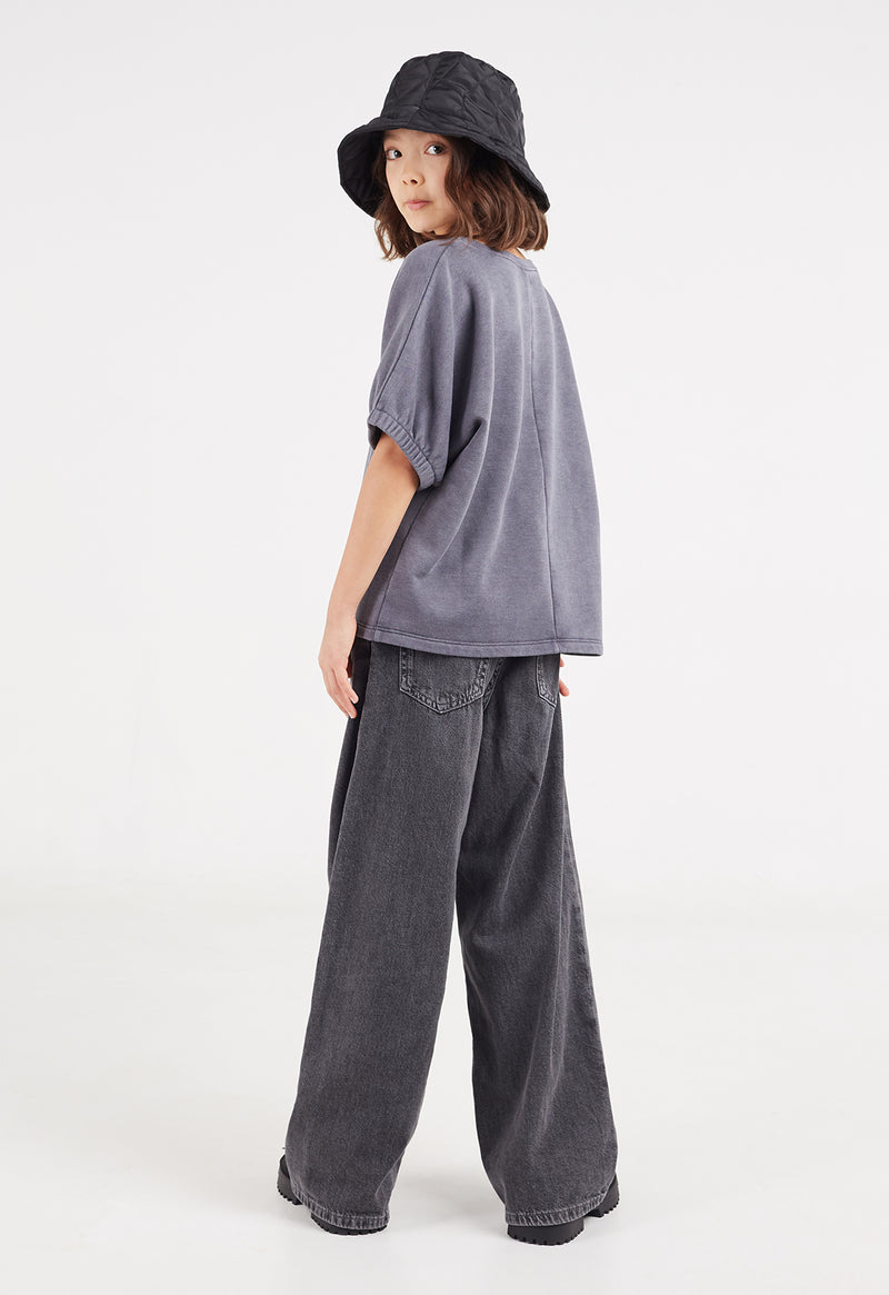 The young girl models the Washed Grey Boxy Girls Sweater T-Shirt by Gen Woo with jeans, sandals and a bucket hat