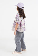 The young girl wears the Pink and Purple Floral Bloom Girls Smock Top by Gen Woo with denim jeans and a baseball hat