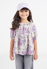 The young girl models the Pink and Purple Floral Bloom Girls Smock Top by Gen Woo with denim jeans and a baseball hat
