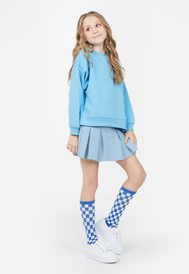 The young girl models the Girls Crew Neck Blue Sweater by Gen Woo with a blue skirt and socks