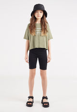 The teen girl wears the Khaki Hashtag Slogan Girls Boxy Cropped T-Shirt by Gen Woo with black bicycle shorts and sandals