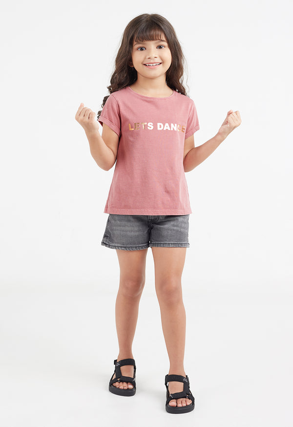 The young girl wears the “Let’s Dance” Girls Gold Foil Slogan T-Shirt by Gen Woo with denim shorts and sandals