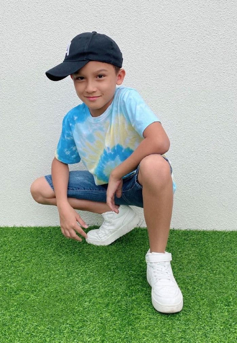 The young boy poses wearing the Boys Blue and Yellow Spiral Tie-Dye T-Shirt by Gen Woo with a baseball hat, denim shorts and sneakers