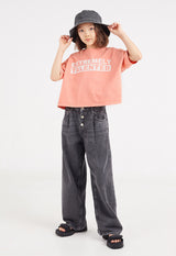 The teen girl wears the Pink “Extremely Talented” Slogan Girls Boxy Cropped T-Shirt by Gen Woo with denim jeans and a bucket hat