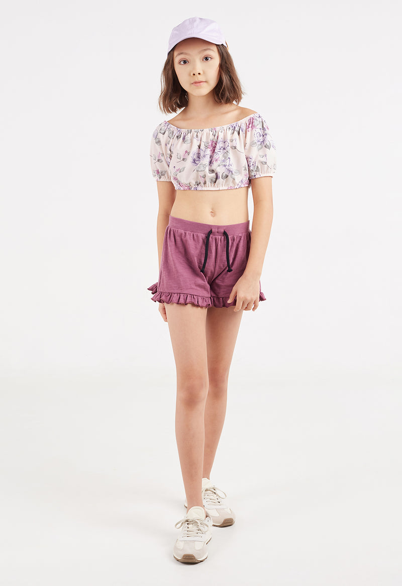 The young girl models the Floral Bloom Girls Crop Top by Gen Woo with shorts, a hat and trainers