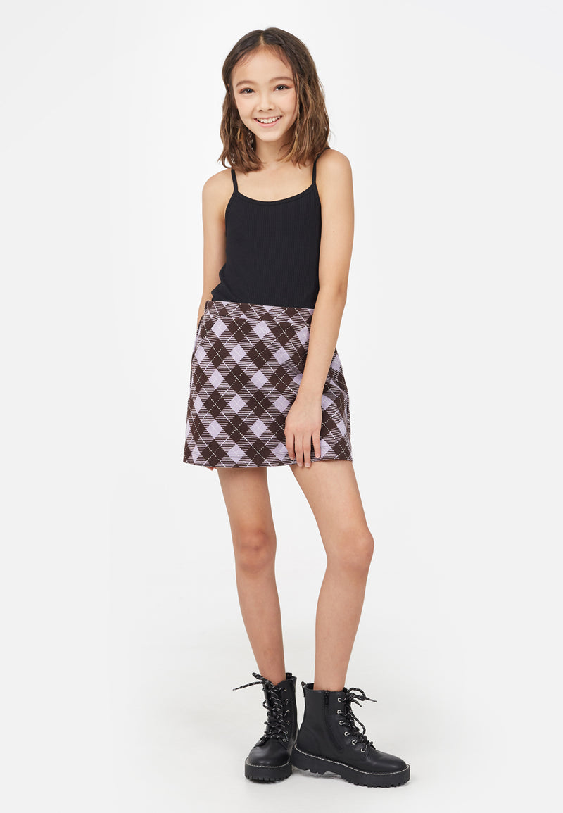 The young girl models the Black Spaghetti Strap Girls Cami Top by Gen Woo with a plaid skirt and chunky boots
