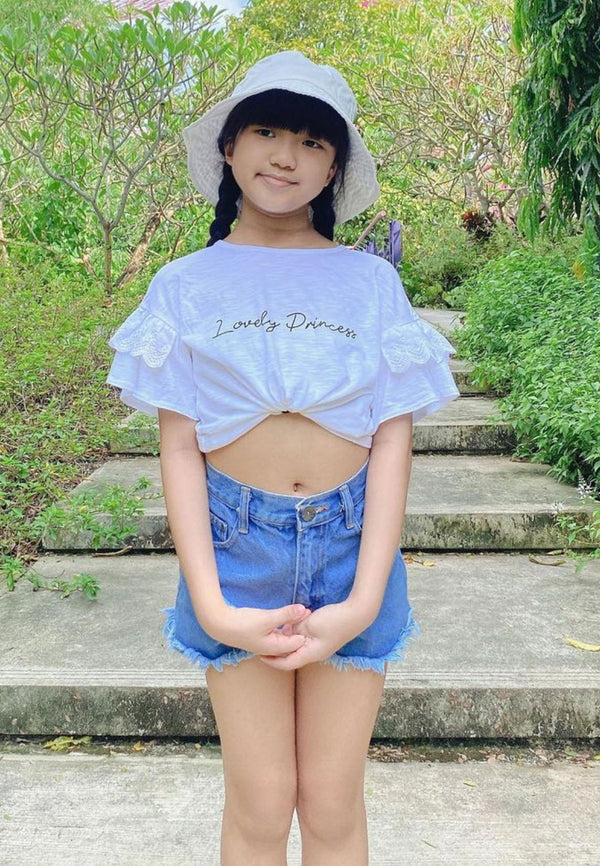 The young girl wears the “Lovely Princess” Broderie Trim Girls Crop Top by Gen Woo with denim shorts